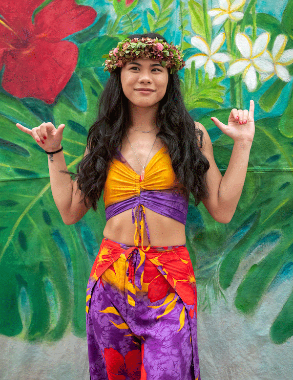 Kayley in front of a tropical themed background showing giving shakas with both hands.