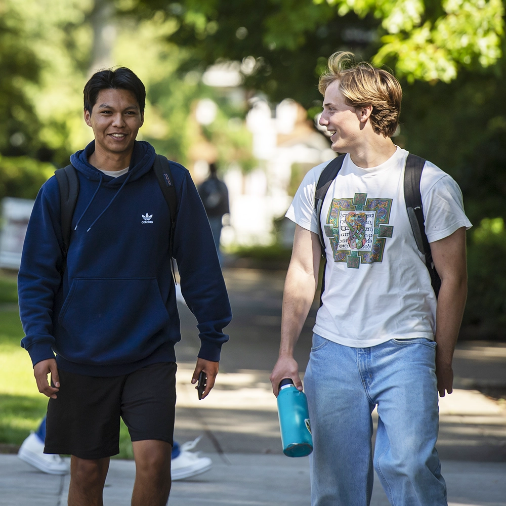 Two students walking on the sidewalk, talking to each other.