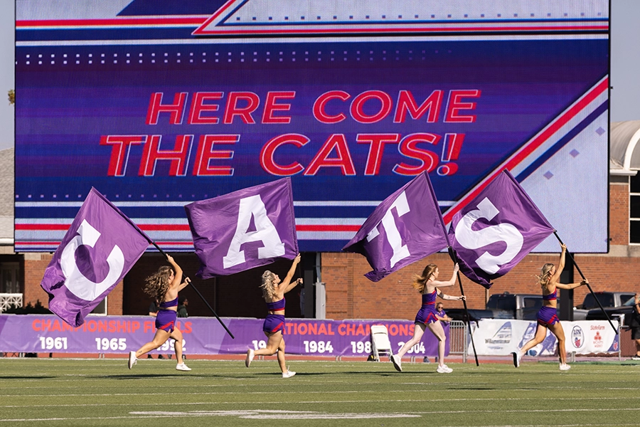 cheerleaders holding flags that spell out "CATS" as they run across the football field.