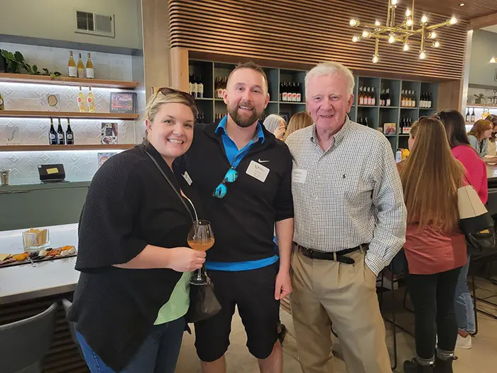 Three smiling people stand next to each other in Troon Vineyard in Downtown McMinnville. They are dressed casually and the mood is happy and relaxed. Behind them is a wine bar and a display of wine while other attendees mill about in the background. 