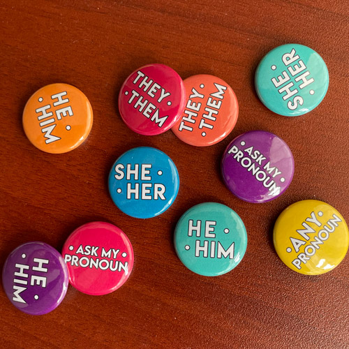 A grouping of colorful pins with different pronouns including they/them, any pronoun, ask my pronouns, she/her, he/him.