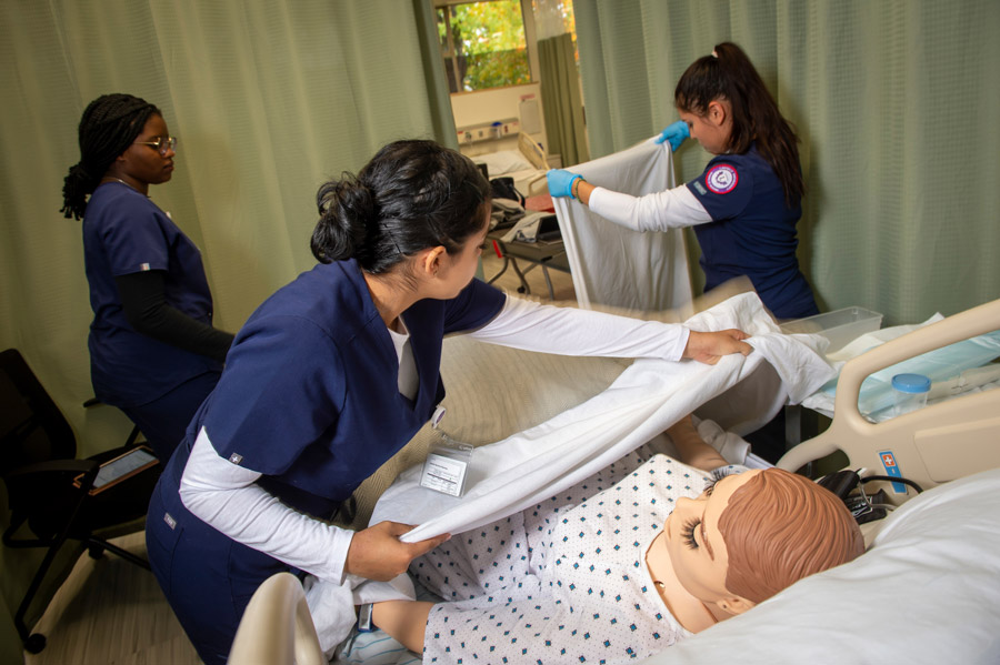 Three students in simulation lab with manikin in hospital bed.