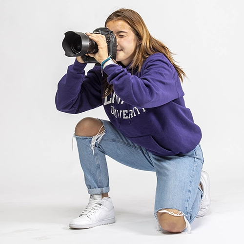 A journalism student looking through the viewfinder of a camera, in a studio with a white background.