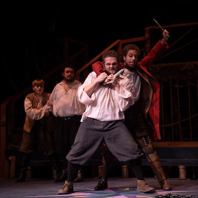 A scene from Treasure Island with Long John Silver (played by Robert Turner '24) holding a knife to Killigrew the Kind's neck (played by Nathan Dillon '20) from behind.