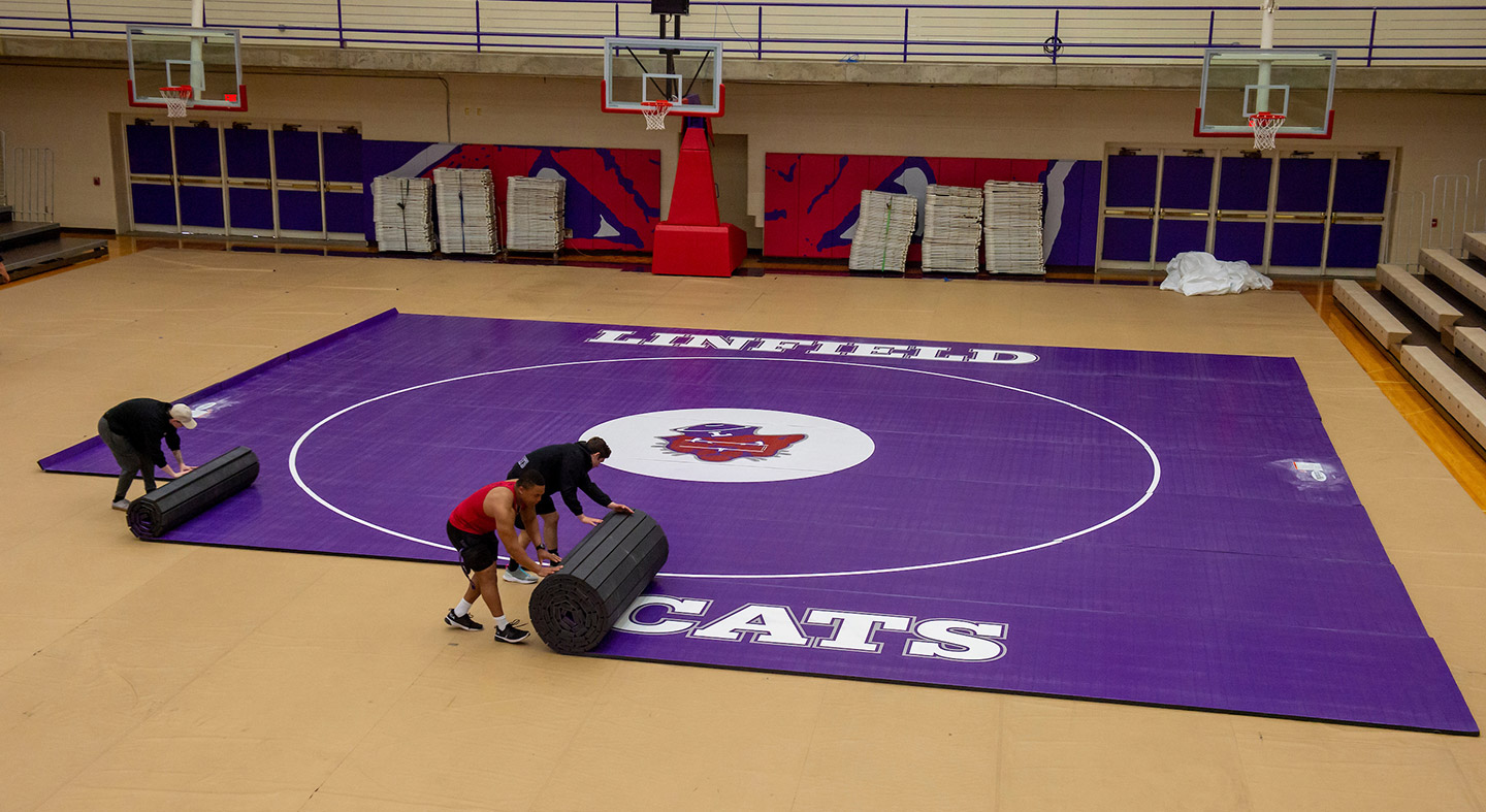 the wrestling mat being rolled out in Ted Wilson Gymnasium by three people.
